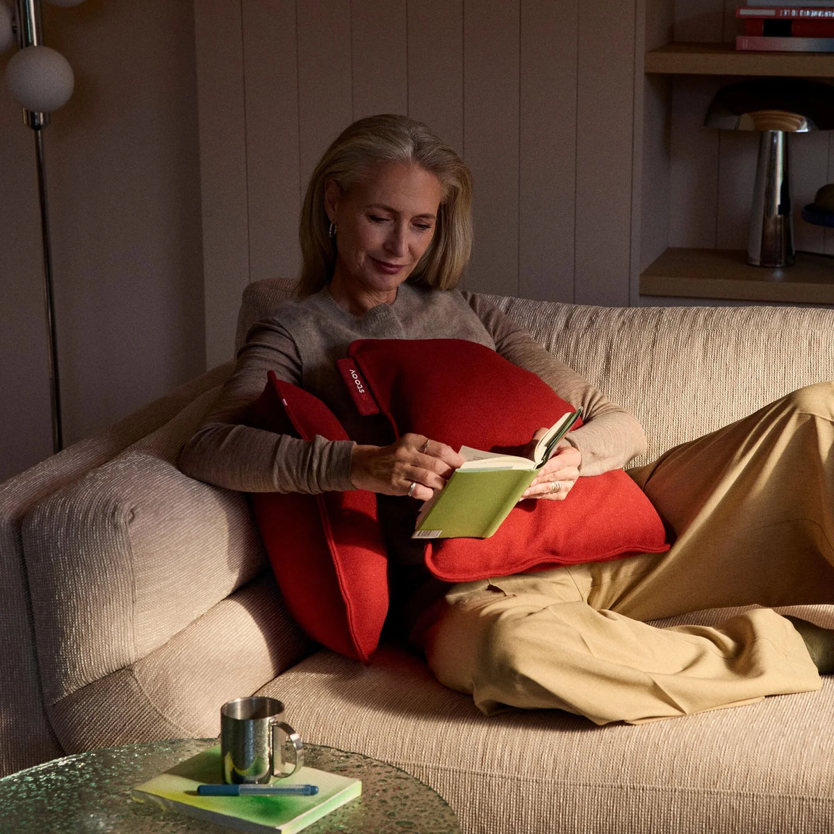 Stoov Ploov Flex warmtekussen 45x60cm in aarde rood, geschikt voor binnengebruik, liggend op een bank met vrouw die leest.